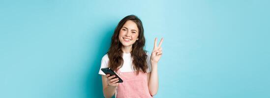 Online shopping. Cute and cheerful young woman smiling, holding smartphone and showing peace v-sign at camera, standing against blue background photo