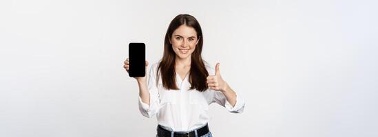 Smiling woman showing thumbs up, recommending app, mobile phone screen, empty interface on smartphone, standing against whte background photo