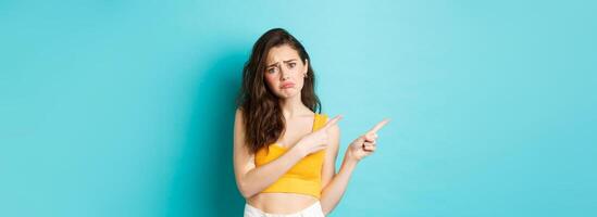 Upset and gloomy brunette girl complaining, pointing fingers left at sad unfair thing, showing bad news, standing against blue background photo