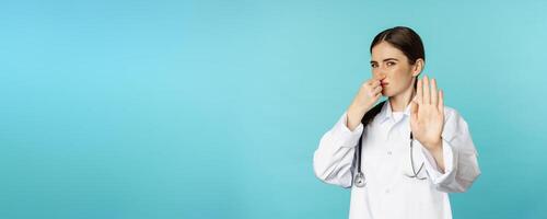 Woman doctor dislike, shut nose from bad smell, reek, showing rejection, stop gesture, standing against torquoise background photo