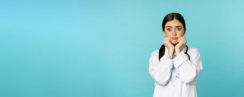 Scared and worried woman doctor in white coat, looking anxious and insecure, shaking from fear, standing over torquoise background photo