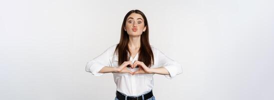 Lovely feminine woman showing heart sign, romantic gesture and smiling with care and tenderness, standing over white background photo