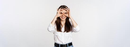 Funny girl looking through hand binoculars, zero gesture, standing over white background and smiling, seeing smth interesting far away, white background photo