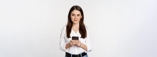 Young woman with smartphone, smiling and looking at camera, using mobile phone app, cellular technology and online shopping concept, white background photo