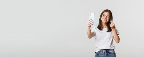 Happy beautiful young woman showing heart gesture and taking selfie on smartphone and smiling photo