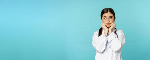 Scared and worried woman doctor in white coat, looking anxious and insecure, shaking from fear, standing over torquoise background photo
