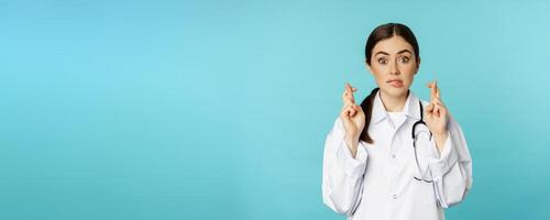 Hopeful woman doctor, lab worker cross fingers and smiling, praying, making wish, anticipating, standing over torquoise background photo