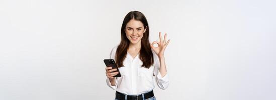 Smiling woman showing okay, ok sign while using mobile phone, approve app on smartphone, excellent service, online store, standing over white background photo