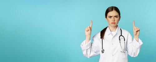 Sad and disappointed healthcare worker, doctor pointing fingers up, frowning upset, showing bad news, standing over torquoise background photo