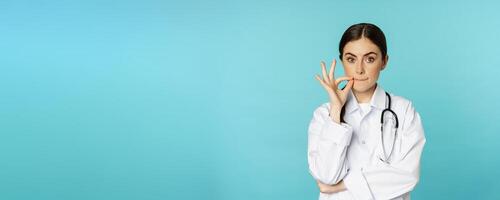 Patient doctor privilege.Young woman healthcare worker showing mouth seal, zipper gesture, standing in white coat against torquoise background photo