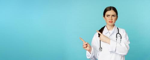 Confused, disappointed medical worker, frowning while pointing fingers left with doubtful, puzzled face expression, standing over torquoise background photo