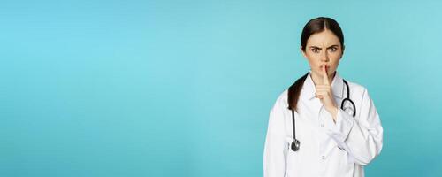 Angry female doctor, healthcare medical worker shushing with disapproval, taboo quiet gesture, silence someone, standing over torquoise background photo