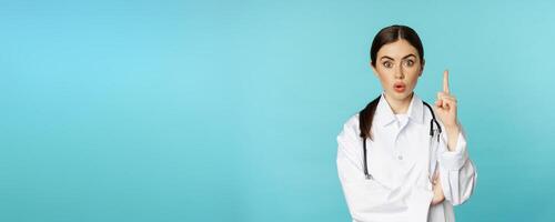 Young doctor, woman physician in white coat raising finger, pointing up, suggesting smth, has solution, revelation, standing over torquoise background photo