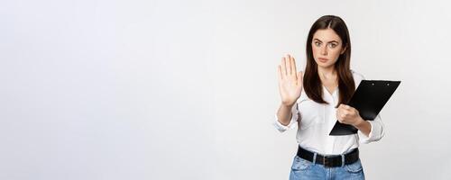 Serious woman with clipboard documents, extending arm, showing taboo stop sign, disapprove, prohibit smth, standing over white background photo