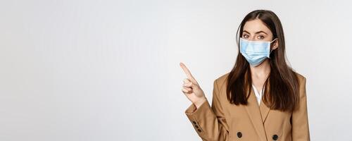 Coronavirus and people concept. Portrait of business woman at workplace wearing face mask, pointing finger left at logo, company banner, white background photo