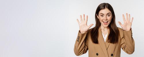 Enthusiastic businesswoman in suit, raising hands palms up, number ten dozen gesture and smiling, standing over white background photo