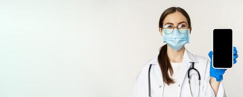 Online medical help concept. Woman doctor in glasses and face mask, showing mobile phone screen, app interface or website for patients, standing over white background photo