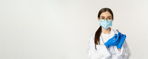 Portrait of young woman, doctor in medical face mask and uniform, showing vaccine, covid-19 vaccination campaign, standing over white background photo