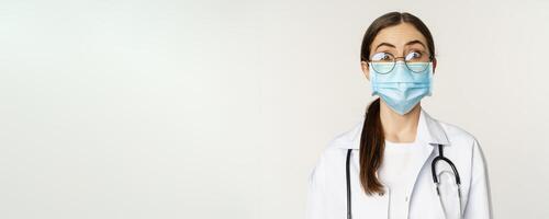 Medical worker, young doctor looking amazed and surprised, wearing face mask to prevent catching covid-19, standing over white background photo