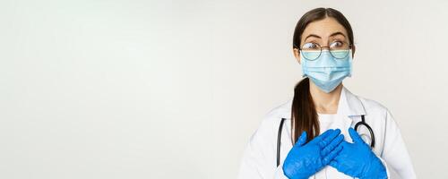 Portrait of surprised and amazed young doctor, looking with fascination, holding hands on heart, standing in medical face mask over white background photo