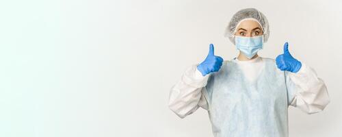 Portrait of doctor in personal protective equipment, showing thumbs up, wearing face mask, concept of covid-19 and pandemic, white background photo