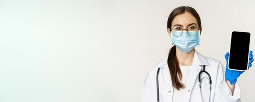Online medical help concept. Woman doctor in glasses and face mask, showing mobile phone screen, app interface or website for patients, standing over white background photo