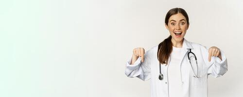 Portrait of happy female doctor, pointing fingers down and smiling, demonstrating promo offer, discount, standing over white background photo