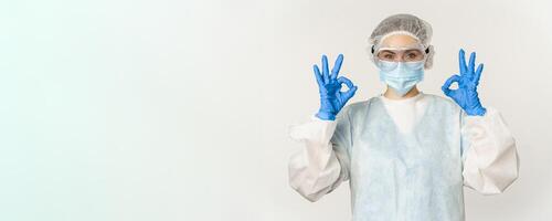 Doctor in personal protective equipment and face mask, showing okay, ok gesture, standing over white background photo