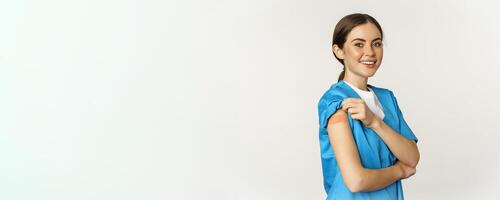 Female nurse, doctor medical worker got vaccinated, showing shoulder with patch after covid-19 vaccine, smiling pleased, standing over white background photo