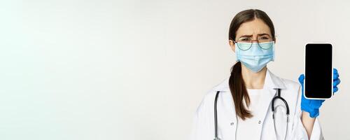 Online medical help concept. Sad and gloomy young doctor in face mask, showing smartphone screen with upset an disappointed expression, white background photo