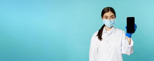 Young woman doctor in medical face mask and hospital uniform, showing mobile phone app, screen interface, online clinic concept, standing over torquoise background photo