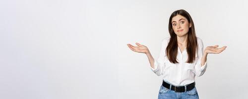 I dont know. Clueless young woman shrugging shoulders and looking unaware, standing over white background photo