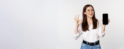 Happy smiling woman with satisfied face, recommending excellent app, showing mobile phone screen, okay sign, standing over white background photo