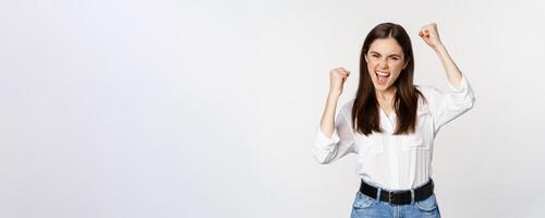 Excited young adult woman reacting to win, surprise news, screaming and cheering, triumphing, achieve goal and celebrating, standing over white background photo