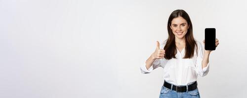 Smiling woman showing thumbs up, recommending app, mobile phone screen, empty interface on smartphone, standing against whte background photo