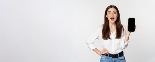 Happy smiling woman showing smartphone app interface, mobile phone screen, demonstrating promo offer, standing over white background photo