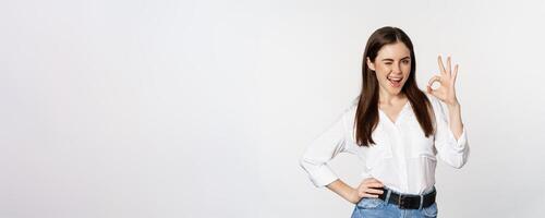 Smiling confident business woman, winking, showing okay ok sign, excellent, no problem gesture, standing over white background photo
