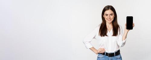 Confident woman in corporate clothes, showing smartphone screen, mobile interface of an application, standing over white background photo