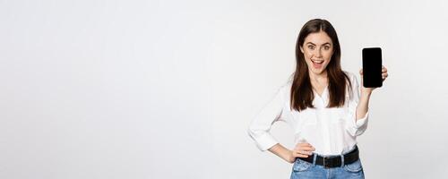 Happy smiling woman showing smartphone app interface, mobile phone screen, demonstrating promo offer, standing over white background photo