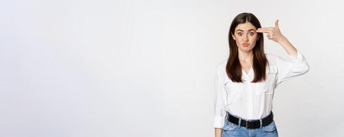 Image of annoyed young woman showing finger gun gesture over head, bothered by smth, tired, standing over white background photo