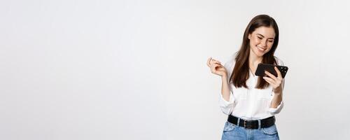 Happy smiling woman watching on mobile phone, looking at smartphone and laughing, standing over white background photo