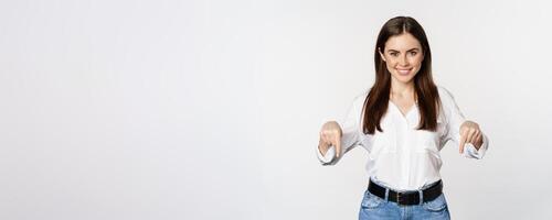 Confident smiling woman pointing fingers down, showing logo, banner below, recommending click on link, standing over white background photo