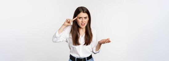Are you dumb. Annoyed woman scolding, roll finger over head and staring at camera, bothered by someone crazy, standing over white background photo