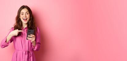 Young woman pointing finger at her phone and smiling, talking about smartphone app, showing something interesting online, standing over pink background photo