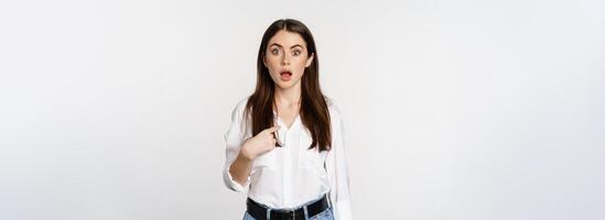 Portrait of young woman pointing at herself with disbelief, surprised face expression, being named or chosen, standing over white background photo