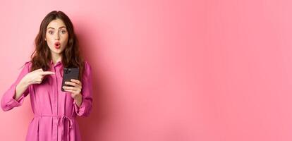 Young woman pointing finger at her phone and gasping wondered, talking about smartphone app, showing something interesting online, standing over pink background photo