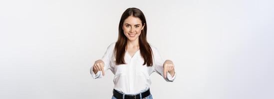 Confident smiling woman pointing fingers down, showing logo, banner below, recommending click on link, standing over white background photo