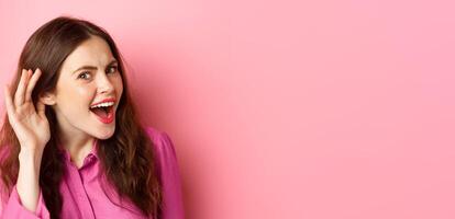 Close up of excited young woman overhear juice rumor, lean closer to camera with hand near ear, eavesdropping, standing over pink background photo