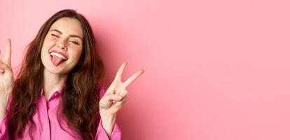 Close up of funny and happy young woman having fun, winking and showing tongue, making v-sign peace gesture, standing over pink background photo