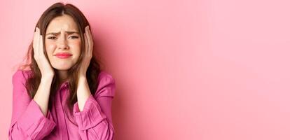 Close up of distressed and bothered young woman covering her ears, grimacing cant bear this anymore, standing overwhelmed against pink background photo